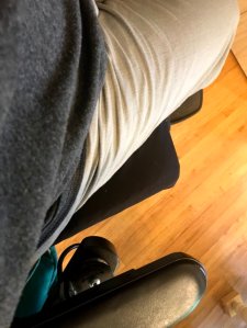 Me in my wheelchair. The only visible parts are pat of the footrest, a corner of the black cushion, part of the right arm rest, a view of the corner of a grey shirt and beige pants.